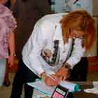 Em um ambiente fechado, algumas pessoas circulam e interagem. A foto destaca mulher que inclina o corpo para frente e escreve num caderno sobre a mesa. Logo atrás dela, outra mulher aguarda. 