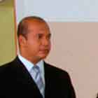 Nesta foto em cores, uma mulher fala ao microfone que um pessoa segura, como se fosse um repórter. Nota-se que ela gesticula ao falar. Ao lado dela, há um homem de terno e uma mulher de vestido vermelho. Atrás deles, percebe-se um banner com o desenho de dois corações sorridentes dentro de uma camisinha.