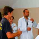 Em um ambiente fechado, algumas pessoas circulam e interagem. A foto destaca mulher de jaleco branco que inclina o corpo para frente e escreve em algo que está sobre a mesa. Logo ao lado dela, outra mulher manuseia alguns papéis, também em cima da mesa.