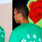 Nesta foto, três rapazes estão de costas para a câmera, em fila para pegar algo sobre a mesa em frente a eles. Nota-se que todos usam camiseta verde com as siglas “SEAP – RJ”. Ao fundo e à direita, percebe-se um banner com o desenho de dois corações sorridentes dentro de uma camisinha.