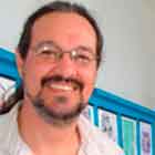 A foto mostra um homem alto, de óculos, barba, bigode e cabelos longos e amarrados ao lado de uma mulher, também alta, usando vestido vermelho. Nota-se que os dois abrem um largo sorriso. Atrás deles, percebe-se uma parede azul onde foram afixadas telas ilustradas com cartuns coloridos.