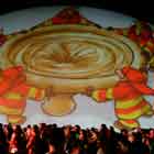 Esta foto colorida revela grande número de pessoas em pé próximas a uma grande cúpula onde foi projeta a imagem de um cartum. O desenho mostra cinco bombeiros que seguram uma enorme camisinha como se fosse uma rede. Eles olham para cima e direcionam o preservativo.