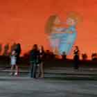 Nesta foto em cores veem-se pessoas em pé e sentadas próximas a uma grande cúpula onde foi projeta a imagem de um cartum. Percebe-se, no desenho, que um casal de mulher e homem se abraça e se beija. Sobre o fundo vermelho, surge um grande coração amarelo, preenchido por incontáveis camisinhas.