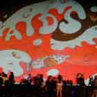 Algumas pessoas em pé e outras sentadas ao redor de uma grande cúpula branca aparecem nesta foto em cores. Percebe-se que a construção recebe em sua superfície a imagem projetada de um cartum. No desenho, notam-se ondas de um mar vermelho povoadas por pessoas que flutuam na água. Observa-se que alguns casais estão abraçados ou se beijando dentro de uma camisinha gigante e lacrada.