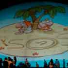 Algumas pessoas em pé e outras sentadas ao redor de uma grande cúpula branca aparecem nesta foto em cores. Percebe-se que a construção recebe em sua superfície a imagem projetada de um cartum. No quadrinho, uma pequena ilha com dois coqueiros abriga um casal de mulher e homem. Nota-se ainda que sobre a areia, escreveram a palavra “SOS”, sendo que o “O” é o desenho de uma camisinha.