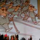 Nesta foto em cores uma superfície arredondada recebe a imagem projetada de um cartum. Percebem-se pessoas em pé e sentadas no chão, em torno da construção. O desenho mostra um homem mal cuidado, sentado no chão e mendigando. Nota-se sobre a cabeça dele uma placa com a sigla “HIV”. Logo antes de cruzar o caminho dele, uma multidão de homens de terno usa também óculos escuros e bengalas, e não dá atenção ao pedinte.