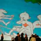 Nesta foto em cores veem-se pessoas em pé e sentadas próximas a uma grande cúpula onde foi projetada a imagem de um cartum. Percebe-se, no desenho, cena em que várias pessoas divertem-se em parque. Nota-se que têm uma cruz vermelha estampada no peito. Além disso, veem-se corações vermelhos na copa de uma árvore, como se fossem maçãs. Percebe-se ainda o céu azul com algumas nuvens.