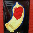 Preso a parede de tijolos vermelhos, há um banner de fundo preto e letras coloridas. Nota-se o desenho de uma camisinha com dois corações sorridentes dentro. Ao final, lê-se:”I Festival Internacional de Humor em DST e Aids”.