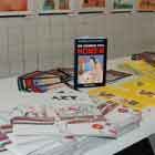 Esta foto colorida mostra, em perspectiva, parede repleta de cartuns. Na mesa que se encontra logo abaixo, há materiais impressos de folheteria, como fôlderes e filipetas.