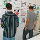 Nesta foto, três pessoas, de costas para a câmera, observam parede repleta de cartuns coloridos. Percebe-se o detalhe da porta de grade ao fundo.