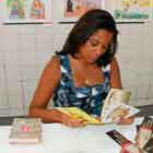 Esta foto mostra, em ângulo fechado, uma mulher sentada a uma mesa sobre a qual há variedade de fôlderes e filipetas coloridas. Na parede branca ao fundo notam-se cartuns afixados.