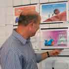 Homem e mulher de perfil para a câmera observam cartuns coloridos afixados em parede branca. Nota-se que o homem aponta para um dos desenhos.