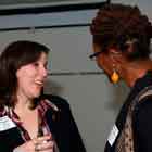 Nesta foto em close e em cores, duas mulheres estão conversando. No rosto daquela à esquerda, nota-se um sorriso aberto. Da mulher à direita não se vê o rosto, pois está de perfil para a câmera. Apenas nota-se que usa um brinco grande e óculos.