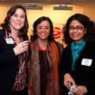 Esta foto em cores mostra três mulheres em pé, lado a lado. Elas olham para a câmera e sorriem. Ao fundo, percebe-se a presença de outras pessoas.