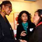 Uma mulher alta, à esquerda, cumprimenta outra, posicionada à direita. Elas se olham e sorriem. Entre as duas, há também outra mulher, que direciona o olhar para aquela à direita. Ao fundo, nota-se parte de um banner afixado à parede. 