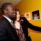 Nesta foto em close, um homem e uma mulher apreciam cartuns afixados em uma parede amarela em frente a eles. Nota-se que o homem deixa escapar um tímido sorriso, enquanto a mulher estende a mão em direção às obras e sorri largamente. 