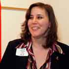 A foto revela mulher em pé, diante de tipo de púlpito. Ela olha levemente para a esquerda e sorri. Ao lado dela, nota-se um banner afixado na parede onde se lê, em Inglês: “1981 first 25 years in the fight. 2006 against Aids