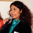 Esta fotografia em cores revela momento em que uma mulher fala alegremente ao microfone, enquanto outra, à esquerda, olha para a discursante com um largo sorriso.