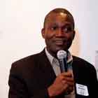 A foto apresenta um homem de terno falando ao microfone. Nota-se no rosto dele uma feição leve, prestes a revelar discreto sorriso. À esquerda, afixado à parede branca, há um banner com letras em destaque dentro de uma faixa vermelha em espiral. É possível ler algumas dessas palavras em Inglês, entre elas: “15 years... against Aids