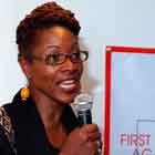 Em close, esta foto revela momento em que uma mulher fala ao microfone. Ao lado dela, em destaque, nota-se um banner afixado à parede onde se lê, em Inglês: “First in the figh agaisnt Aids. GMHC.