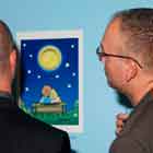 A foto em close, mostra dois homens de costas observando cartum afixado na parede em frente a eles. Percebe-se que a obra que apreciam revela um casal sentado em um banco, olhando para o céu estrelado onde a lua, na verdade, é uma camisinha.