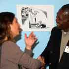 A foto colorida mostra, à esquerda, mulher cumprimentando homem sorridente ao centro. À direita, nota-se outra mulher que observa a conversa.