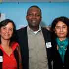 A foto em cores mostra grupo de pessoas em pé, bem próximas umas às outras. Nota-se que olham para a câmera e algumas sorriem. Percebe-se ainda que há somente um homem ao centro, em meio a quatro mulheres.