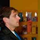 Esta foto em close revela homem de perfil, com feição séria, observando cartum azul afixado na parede amarela em frente a ele. Ao fundo, notam-se algumas publicações à mostra.