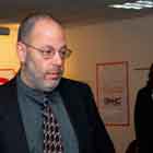 Nesta foto em cores, um homem conversa com uma mulher enquanto gesticula. Nota-se a feição séria dele, olhando para sua interlocutora. Ao fundo, observam-se pessoas circulando pelo espaço onde se encontram.