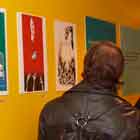 Nesta foto, uma parede amarela serve de fundo para vários cartuns coloridos. Notam-se algumas pessoas apreciando as obras expostas.