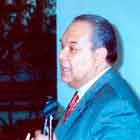 Nesta foto há um senhor de terno falando ao microfone. Em frente a ele há um pequeno balcão com algumas publicações. À esquerda, nota-se uma mulher que observa o discursante. Já ao fundo da imagem, percebe-se um grande painel com cartum colorido. 