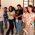 Esta foto em cores mostra grupo de pessoas olhando para frente, onde alguma coisa acontece. Nota-se que algumas pessoas têm um papel nas mãos. Ao fundo, percebe-se a copa de uma árvore do lado de fora da janela.