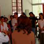 Pessoas em pé, olhando para a esquerda aparecem nesta foto em cores. Nota-se ao fundo uma grande estrutura vertical vermelha que abriga um dos painéis da mostra. Percebe-se também que a foto foi tirada em um local amplo e fechado. 