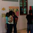 Nesta foto, um senhor à esquerda observa cartuns afixados na parede. Logo após a janela que se encontra ao lado do homem, há um pequeno grupo de pessoas que também observam as obras expostas.