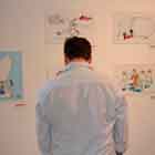 Em pé, de costas para a câmera, notam-se um homem e uma mulher observando os cartuns afixados na parede.