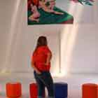 Esta foto mostra, à esquerda, uma estrutura vertical vermelha onde se encontram dois cartuns. Ao fundo, duas mulheres observam as peças da mostra. Na parede alta da sala fechada nota-se um grande cartum colorido. O desenho revela homem nu deitado na cama, acompanhado de uma mulher, também nua. Nota-se que ele estica o braço e o dedo indicador. Ao mesmo tempo, um homem de cabelos e barba compridos e brancos, cercado por anjos, estica a mão em direção ao homem e oferece-lhe uma camisinha.