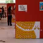 Em um local amplo e fechado avista-se uma exposição de cartuns. Observam-se estruturas verticais vermelhas que funcionam como suporte para os desenhos. Entre as obras, duas se destacam. A primeira, à esquerda, mostra homem seminu segurando nota de dinheiro. Percebe-se a que ele tem dúvida ao olhar um pão e uma embalagem de camisinha. O segundo cartum revela dezenas de pintinhos amarelos separados por uma linha pontilhada vermelha. Os pontos levam a um casal de pintinhos cobertos por camisinhas. Notam-se pequenos corações vermelhos flutuando sobre a dupla.