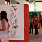 Esta foto revela pessoas visitando exposição de cartuns. Nota-se que as obras foram afixadas em estruturas verticais vermelhas. Uma delas, à esquerda, mostra desenho em preto e branco de homem com dinheiro na mão e expressando dúvida ao olhar para baixo.