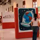 Esta foto revela duas meninas visitando exposição de cartuns. Nota-se que as obras foram afixadas em estruturas verticais vermelhas.