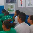 Nesta foto, há pessoas sentadas de costas para a câmera, olhando para frente na sala onde estão. Percebe-se, na parede à direita, um banner preenchido com alguns cartuns coloridos.