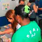 Uma mulher com camiseta verde escrito “SEAP – RJ” está de costas nesta foto. Ela aguarda moça que pega alguns itens de folheteria expostos sobre mesa. Nota-se, ao fundo, um banner afixado na parede e preenchido com alguns cartuns. No canto direito da imagem, identifica-se um grupo de quatro pessoas conversando.