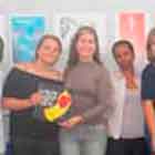 Nesta foto em cores, um grupo de pessoas posicionadas lado a lado posa para a câmera. Nota-se que estão em uma sala fechada, na frente de um grande banner preenchido com cartuns. Algumas pessoas sorriem e uma das mulheres mostra exemplar da publicação  Festival Internacional de Humor em DST e Aids.