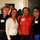 Quatro mulheres aparecem nesta foto lado a lado, posando para a câmera. Somente a da direita expressa uma feição séria, as outras sorriem. Ao fundo, notam-se algumas pessoas em frente a painéis que expõem cartuns coloridos.