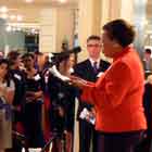 Nesta foto em cores, nota-se mulher sobre um degrau, falando ao microfone. Ao redor dela, há dezenas de pessoas observando-a e fotografando-a.