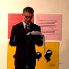 Esta foto revela homem de terno falando ao microfone sobre um pequeno tablado. Ao fundo, notam-se cartuns coloridos afixados na parede branca.