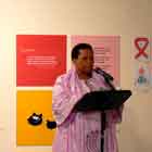 Esta foto revela mulher falando ao microfone sobre um pequeno tablado. Ao fundo, notam-se cartuns coloridos afixados na parede branca e um senhor em pé, lendo papel em suas mãos.