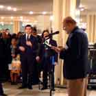 Nesta foto em cores, nota-se senhor sobre pequeno tablado, falando ao microfone. Ao redor dele, há dezenas de pessoas observando-o.