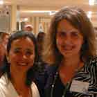 Duas mulheres aparecem nesta foto lado a lado, posando para a câmera. Nota-se que as duas sorriem e a mulher à direita segura uma taça com líquido colorido. Ao fundo, notam-se algumas pessoas circulando pela mostra.