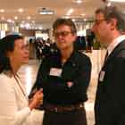 Três pessoas aparecem em pé, conversando nesta foto em cores. Notam-se algumas pessoas espalhadas pelo salão onde se encontram.