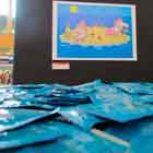 Esta foto em cores mostra várias embalagens azuis de camisinha bem próximas à câmera. Ao fundo, colados em suporte vertical preto, notam-se dois cartuns. Observa-se que um deles mostra dois homens num pequeno pedaço de areia cercado pelo mar.